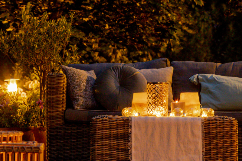 Burning Candles Indoors On A Cozy Patio At Night With Lanterns And Glowing Candles On A Wicker Table Surrounded By Plants, Pillows, And Soft Lighting.