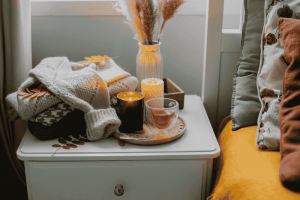 blog 2 A Cozy Autumn Setup With A Burning Soy Candle, Tea, And Knit Blankets On A Bedside Table.